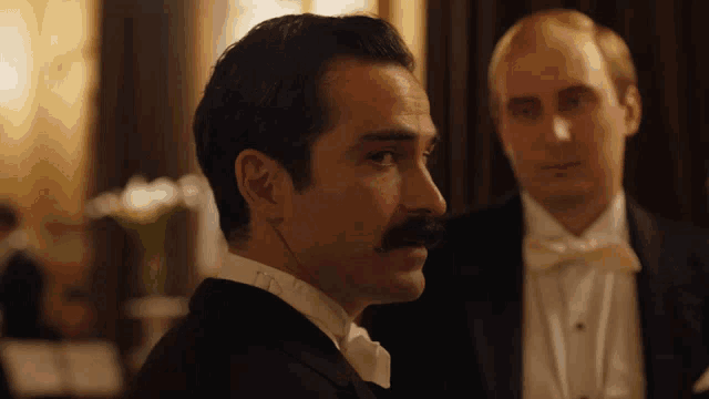 a man with a mustache wearing a tuxedo and bow tie stands next to another man