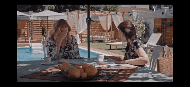 two women sit at a table with a bowl of oranges and a cup of coffee