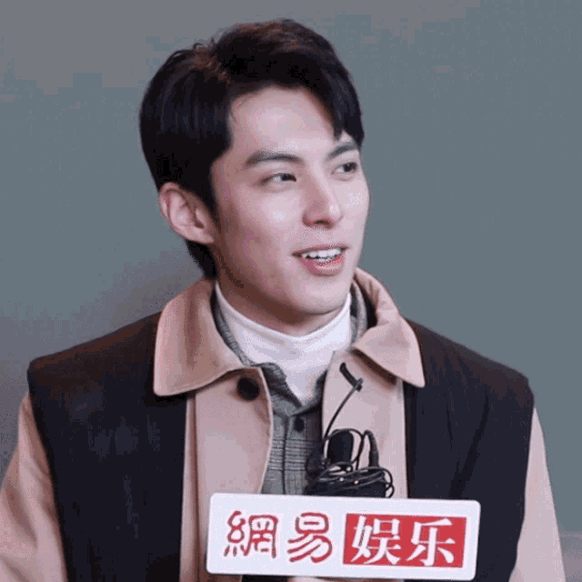 a man wearing a tan jacket and a black vest is smiling and holding a sign that says " 網易 娱乐 "