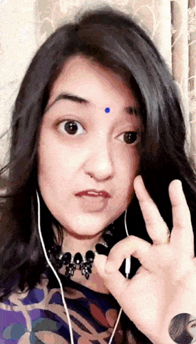 a woman with a blue dot on her forehead is wearing ear buds and making an ok sign