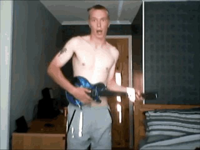 a shirtless man is holding a blue guitar in a bedroom
