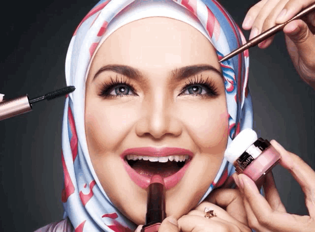a woman wearing a hijab is getting her makeup done by a woman holding a bottle of l' oreal