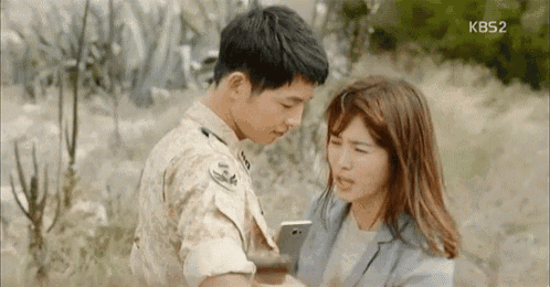 a man and a woman are standing next to each other in a field . the woman is holding a cell phone .
