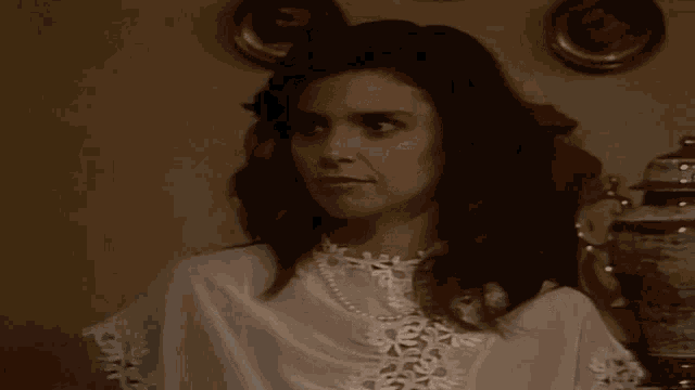 a woman wearing a white top and a pearl necklace looks at the camera
