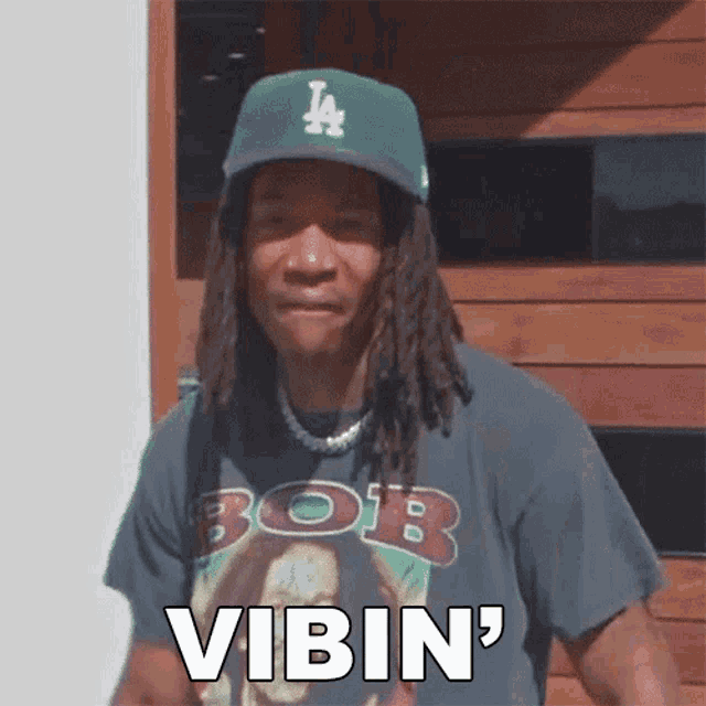 a man with dreadlocks wearing a la hat and a bob shirt says vibin