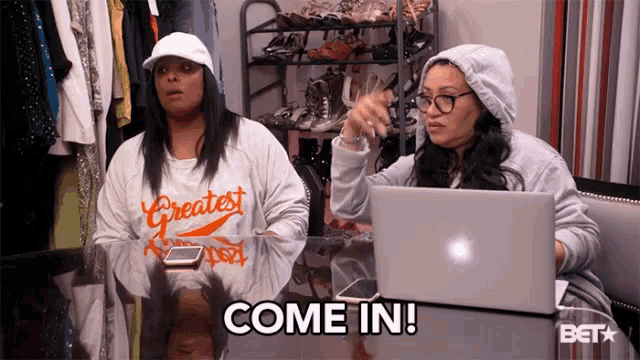 two women are sitting at a table with a laptop and one of them is wearing a shirt that says " greatest "