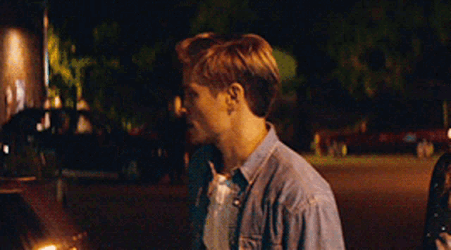 a young man in a denim shirt is walking down a street at night
