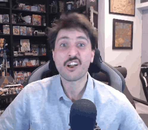 a man with a mustache is talking into a microphone in front of a bookshelf