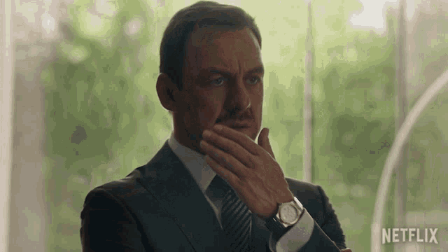 a man in a suit and tie is covering his mouth with his hand and a netflix logo is visible behind him