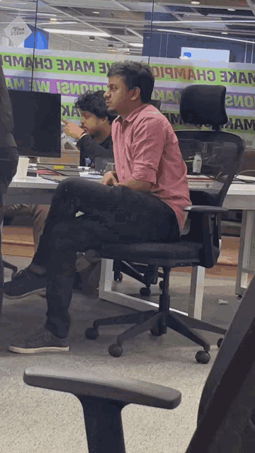 a man in a pink shirt sits in a chair in front of a sign that says ' oi9man '