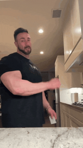a man in a black t-shirt with the word muscles on it is standing in a kitchen