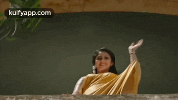 the woman is wearing a yellow saree and waving her hand .