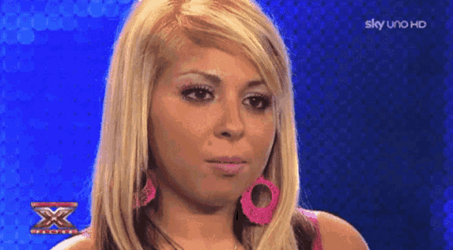 a woman with blonde hair and pink earrings looks at the camera with a sky uno hd logo behind her