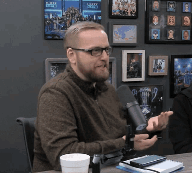 a man wearing glasses talks into a microphone in front of a wall that has pictures on it that say hirez expo