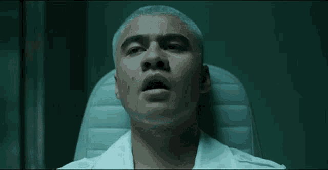 a man in a white shirt is sitting in a chair with a hand covering his mouth .