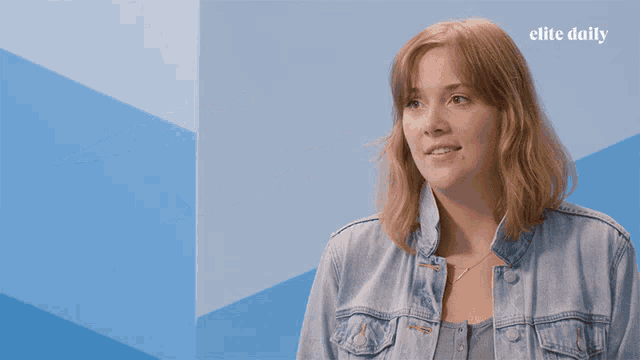 a woman in a denim jacket stands in front of a blue wall with the elite daily logo