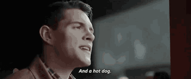 a man is sitting in a dark room and talking about a hot dog .