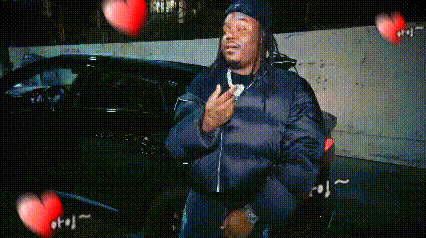 a man in a black jacket is sitting in front of a car with hearts flying around him .