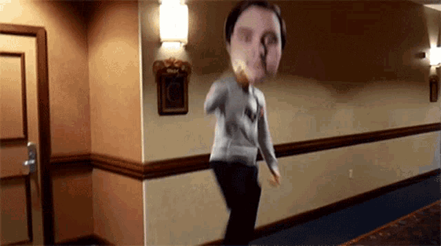 a man in a grey sweater is walking down a hallway with a picture of his face on the wall behind him