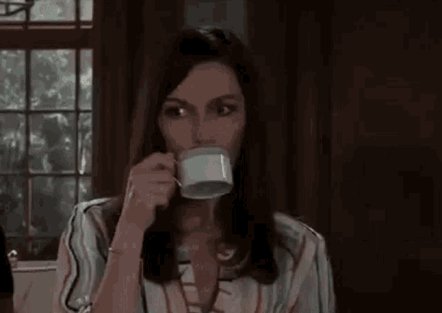 a woman is drinking a cup of coffee while sitting in front of a window .