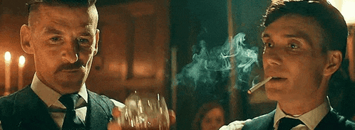 two men in suits and ties are smoking cigarettes and drinking whiskey .