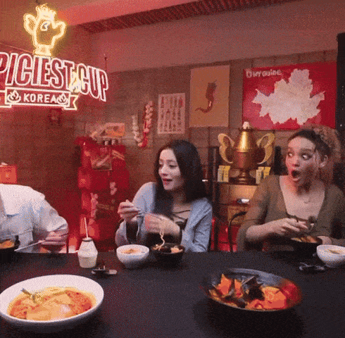 three people are sitting at a table with bowls of food in front of a sign that says ' piciest cup korea '