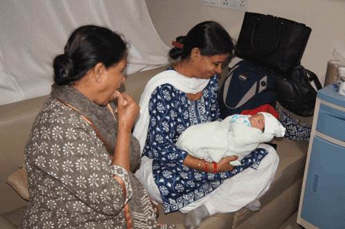 a woman sitting on a couch holding a baby and talking to another woman