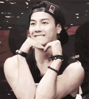 a young man wearing a hat and bracelets is smiling with his hands on his chin .