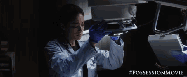 a woman in a lab coat and blue gloves is looking at something in a dark room with the hashtag #possessionmovie