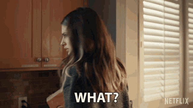 a woman is standing in a kitchen with shutters and says " what "