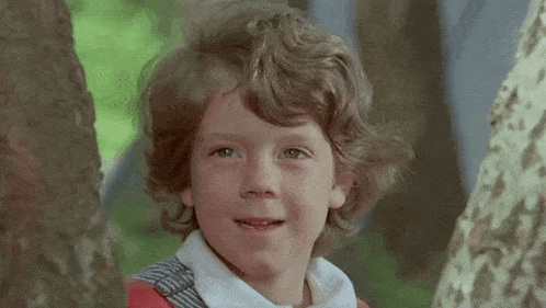a young boy is peeking out from behind a tree giving a thumbs up .