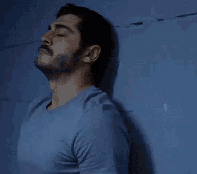 a man with a beard and mustache is leaning against a blue wall with his eyes closed .
