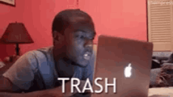 a man is laying on a bed looking at a laptop with the word trash on it .