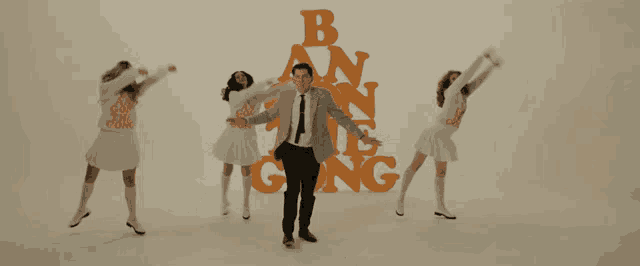 a man in a suit and tie stands in front of a group of women dancing in front of the letters b and n