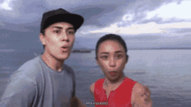 a man and a woman are standing in front of a body of water . the man is wearing a hat .