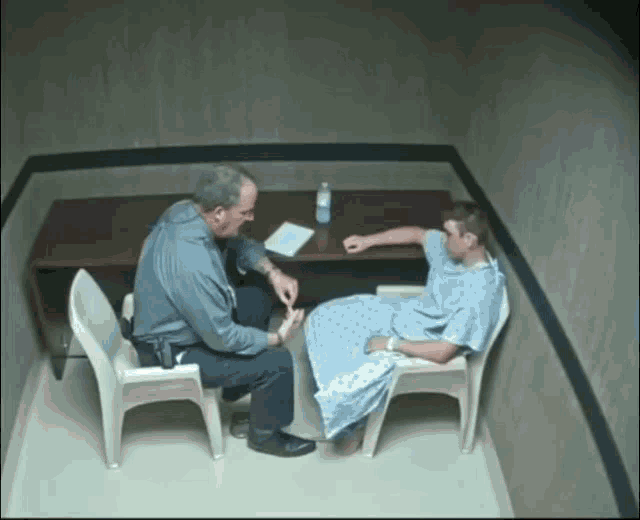 a man in a hospital gown sits in a chair while a police officer talks to him