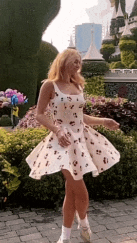 a woman in a white dress with cherries on it is standing on a brick sidewalk .
