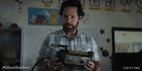 a man in a plaid shirt is holding a ghostbusters device in a classroom .