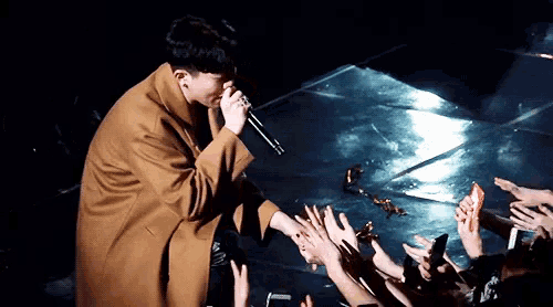 a man in a brown coat is singing into a microphone while a crowd holds their hands up