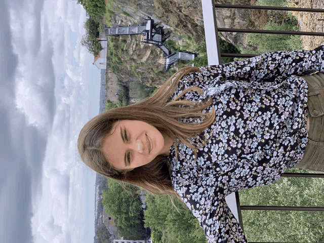 a woman in a floral shirt stands on a balcony overlooking a hillside