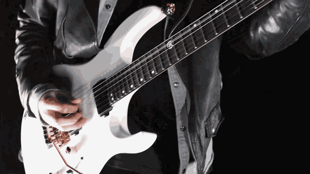 a man in a leather jacket is playing a white electric guitar with the letter t on the neck