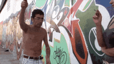 a man without a shirt stands in front of a wall with graffiti on it and a woman smoking a cigarette