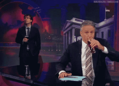 a man in a suit and tie is smoking a cigarette while another man holds a glass of wine