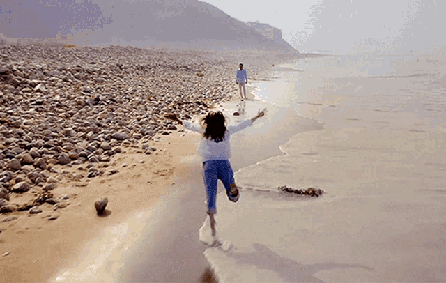 a girl is running on a rocky beach while a man stands behind her