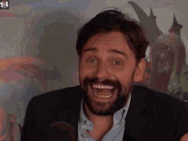 a man with a beard is smiling in front of a microphone with a purple t on the bottom right