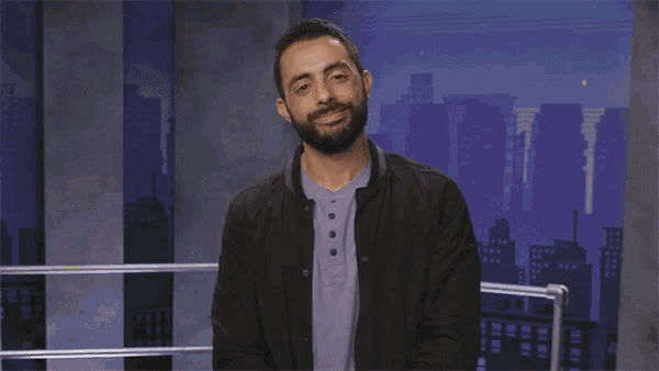 a man with a beard is sitting in front of a city skyline and says `` also fake '' .