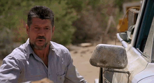 a man with a beard is standing next to a truck in the dirt