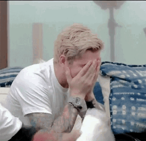 a man in a white shirt is covering his face with his hands while sitting on a bed .