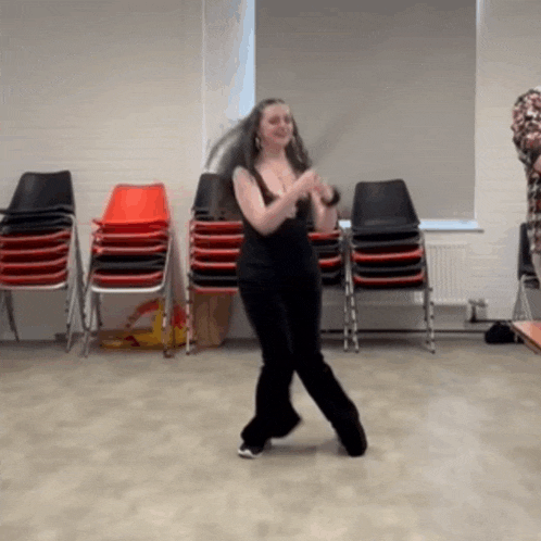 a woman in black pants is dancing in a room with stacks of chairs .