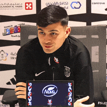 a man is sitting in front of a microphone with a nova logo on the screen